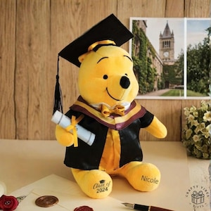 Puede incluir: Un peluche amarillo de Winnie the Pooh con birrete y toga de graduación, sosteniendo un diploma. El peluche tiene impreso "Class of 2024" y "Nicole". El fondo incluye una superficie de madera y una foto enmarcada.