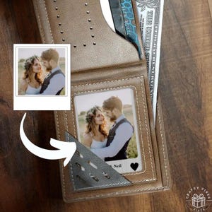May include: A brown leather wallet with a photograph of a couple. The wallet features card slots and a cash compartment. The photograph shows a man and woman in a field. The wallet is on a wooden surface.