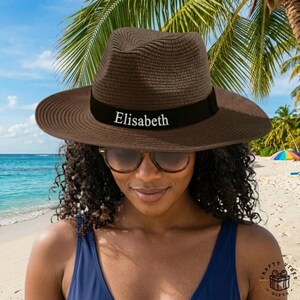 Könnte beinhalten: Brauner Fedora-Hut mit einem schwarzen Band mit dem Namen "Elisabeth" in Weiß. Der Hut hat eine breite Krempe und wird von einer Person getragen, die eine Sonnenbrille und einen blauen Badeanzug trägt. Der Hintergrund zeigt eine Strandszene mit Palmen und einem bunten Regenschirm.