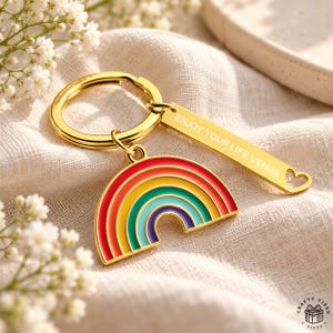 Könnte beinhalten: Ein goldfarbener Metallschlüsselanhänger mit einem halbkreisförmigen Regenbogen-Anhänger mit roten, orangen, gelben, grünen, blauen und violetten Emaille-Streifen. Daran befestigt ist ein rechteckiges goldenes Schild mit der Gravur ENJOY YOUR LIFE VENUS.