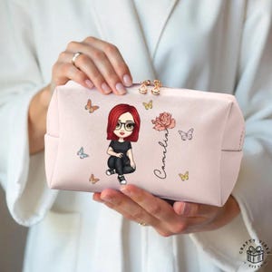 Op de afbeelding: Een lichtroze cosmetische tas met een cartoonillustratie van een persoon met rood haar, een bril en zwarte kleding. De tas heeft vlinder- en rozenontwerpen, met de naam "Camelia" erop geschreven. De tas wordt door een persoon vastgehouden.