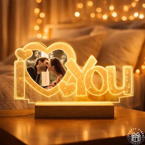 Könnte beinhalten: Ein beleuchtetes "I Love You"-Schild mit einem herzförmigen Rahmen, der ein Foto eines sich küssenden Paares enthält. Das Schild leuchtet warmweiß und steht auf einem Holzsockel. Der Hintergrund ist mit weichem Licht verschwommen.