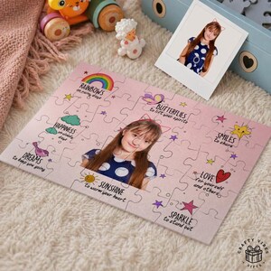 May include: A personalised jigsaw puzzle with a photo of a girl in a blue polka dot dress. The pink puzzle has inspirational words and drawings like rainbows, butterflies, and hearts. A polaroid photo of the girl is also present.