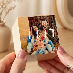 May include: A cube-shaped wooden digital clock featuring a custom printed photograph of a family of three on its front face. The clock displays the time in bright white digital numerals directly over the image of the two adults and one child.