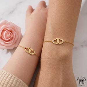 May include: Gold-colored bracelets with interlocking heart pendants. Each heart is engraved with a name and adorned with a small gemstone. The bracelets are displayed on wrists, suggesting a personalized jewelry item.