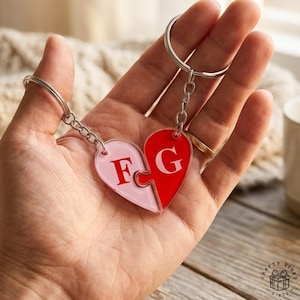 May include: A two-piece heart-shaped keychain with interlocking puzzle pieces. One half is light pink with the letter "F", the other is red with the letter "G". Both pieces are attached to a silver keyring. The keychain is held in a hand.