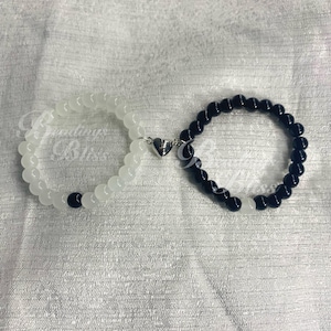 May include: Two beaded bracelets, one white and one black, connected by a silver heart-shaped clasp. The bracelets are made of round beads. The white bracelet has a single black bead, and the black bracelet has a single white bead.