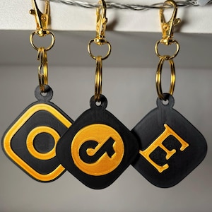 Könnte beinhalten: Drei schwarze Schlüsselanhänger mit goldfarbenen Logos. Die Logos sind ein Kreis mit einem Strich durch, ein stilisierter Buchstabe "C" und ein stilisierter Buchstabe "E".