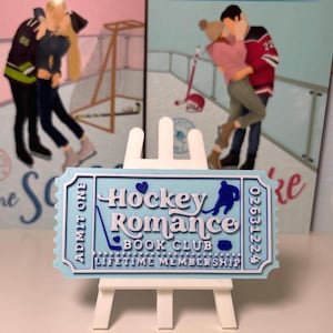 Könnte beinhalten: Ein hellblaues, ticketförmiges Schild mit der Aufschrift "Hockey Romance Book Club Lifetime Membership" wird auf einer kleinen weißen Staffelei präsentiert. Das Schild zeigt eine Eishockeyspieler-Silhouette und eine Hockeyschläger-Grafik. Zwei Bücher von Elle Kennedy sind im Hintergrund.