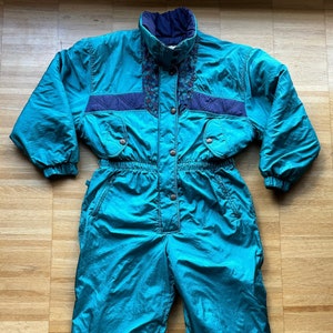 May include: A teal blue ski suit with a navy blue and multi-coloured patterned design on the chest and shoulders. The suit has a zip closure and a snap button closure.