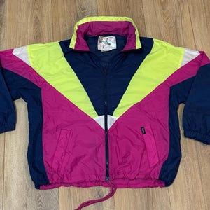 May include: A retro windbreaker jacket with a bold colour block pattern. The jacket combines navy blue, hot pink, white, and neon yellow. It has a full zip, pockets, and a drawstring hem. The collar is a contrasting pink and yellow.