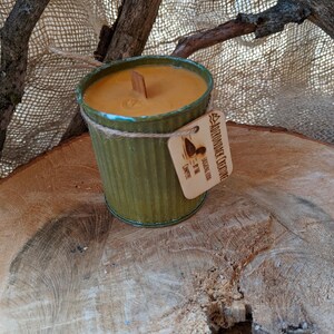 以下が含まれることがあります： 木製の芯が付いた緑色の金属容器に入った手作りのキャンドル。キャンドルは薄茶色のワックスで、「Adirondack Creations」と書かれたタグが付いています。キャンドルは木の輪切りに置かれています。