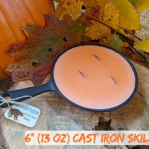 May include: A cast iron skillet candle with an orange wax and three wooden wicks. The candle is 6 inches in diameter and weighs 13 ounces. The candle is on a wooden surface with fall foliage and a pumpkin in the background.