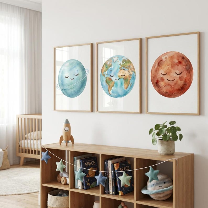 May include: Three framed watercolor prints of planets: Saturn, Earth, and Mars. Each planet has a smiling face. The prints are in wooden frames and hang above a wooden bookshelf with space-themed decor, including a rocket and star garland.
