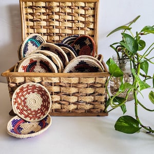 Può includere: Un cesto di vimini intrecciato con un coperchio, pieno di cesti intrecciati colorati. Il cesto è appoggiato su una superficie bianca accanto a una pianta in vaso.