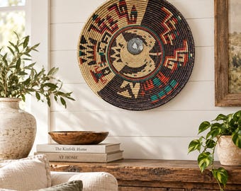 May include: A round wall-mounted woven basket featuring a geometric pattern in black, tan, red, and teal. The center of the basket displays a small circular metallic accent. The design incorporates intricate zig-zag and block motifs.