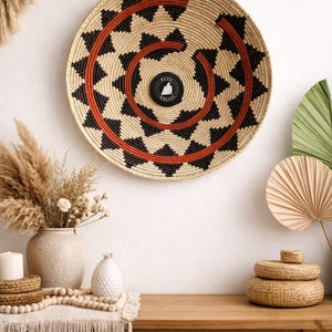 Può includere: Un cesto da parete rotondo intrecciato con un motivo a spirale nero e rosso. Il cesto è appeso a una parete bianca. Un piccolo cerchio nero con testo bianco è al centro. Il cesto fa parte di un tema di arredamento per la casa.
