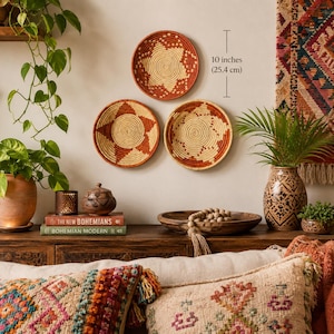 May include: Three round woven wall baskets featuring geometric patterns in terracotta and cream colors. Each basket measures 10 inches in diameter. The baskets are displayed on a wall above a wooden surface with various decorative items.