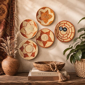 Könnte beinhalten: Fünf runde, geflochtene Wandkörbe mit geometrischen Mustern in Creme und gebranntem Orange. Das Set enthält eine große Holzschale, eine Terrakottavase mit eingravierten Details und zwei gestapelte Bücher mit den Titeln The Kinfolk Home und Habitat.