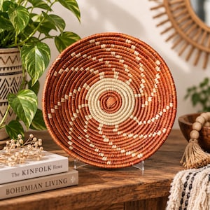Könnte beinhalten: Ein runder, handgeflochtener Wandkorb mit einem Spiralmuster in abwechselnden Farbtönen von tiefem Terrakottabraun und Creme. Das gewickelte Design strahlt von der Mitte aus und erzeugt eine strukturierte, kreisförmige Oberfläche mit Kontrast.