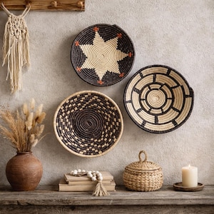 Pode incluir: Três cestos decorativos de parede tecidos em tons de preto e naturais pendurados numa parede cinza claro. Um design de estrela, círculos concêntricos e padrões geométricos adornam os cestos. Um pequeno cesto tecido com tampa, um vaso e uma vela estão numa prateleira de madeira.