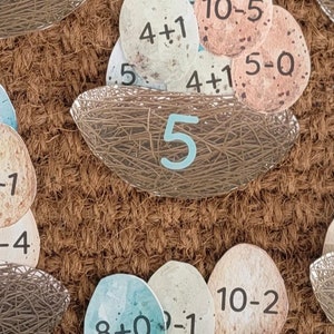 May include: A set of colourful paper eggs with maths problems written on them. The eggs are arranged in nests with a number at the bottom of each nest. The numbers are 5, 6, and 7. The maths problems include addition, subtraction, and multiplication.