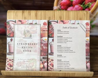 Puede incluir: Un soporte de libro de recetas de madera que muestra un libro de recetas de fresa y una tabla de contenidos. La portada del libro dice "Strawberry Recipe Book" con 16 recetas. La tabla de contenidos enumera bebidas, desayuno, aperitivos, untables y postres.