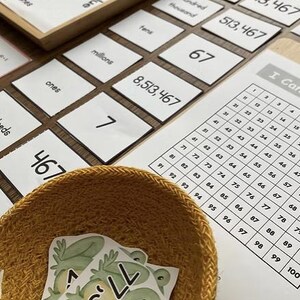 Op de afbeelding: Educatief wiskundemateriaal. Een houten dienblad met plaatswaarde labels en nummerkaarten. Een geweven mand bevat kikker-thema nummerkaarten. Een 100-tabel is ook zichtbaar.
