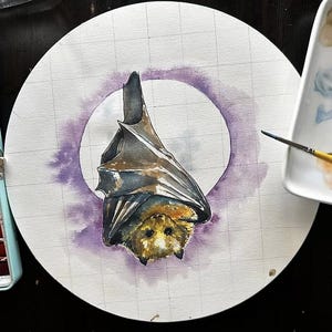May include: Watercolor painting of a bat hanging upside down, centered on a white circle with a purple halo. The bat has brown and black fur with yellow eyes. A watercolor palette and paintbrushes are in the background.
