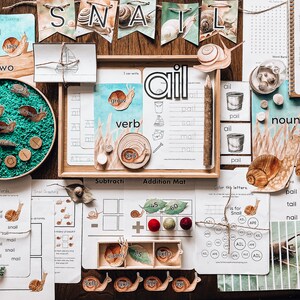May include: Educational snail-themed learning materials. Includes flashcards, wooden snail cutouts, and a tray with green filler. The word "SNAIL" is displayed on a banner. Other cards show words like "verb" and "noun".