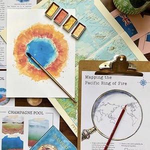 May include: An artistic still life featuring watercolor paints, a paintbrush, and a watercolor painting of a geothermal pool. A clipboard with the text "Mapping the Pacific Ring of Fire" and a passport are also visible.