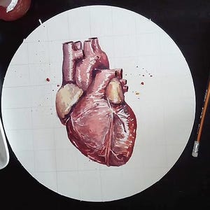 May include: Watercolor painting of a human heart in shades of red and pink, centered on a white circle with a grid pattern. Paintbrushes, a ruler, and a watercolor palette are visible on a black surface.