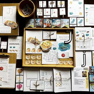 以下が含まれることがあります： 日本のテーマの教育資料のコレクション。フラッシュカード、ワークシート、日本の食べ物と日本の国旗のイラストが描かれた木製トレイが含まれています。資料は、暗い木製の表面に配置されています。