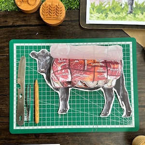 Pode incluir: Um conjunto educativo em madeira com um diagrama de anatomia Angus, um diagrama de vaca com cortes de carne e carimbos de madeira com um design de trator e trigo. Inclui uma bandeja de madeira com a palavra "ANGUS".