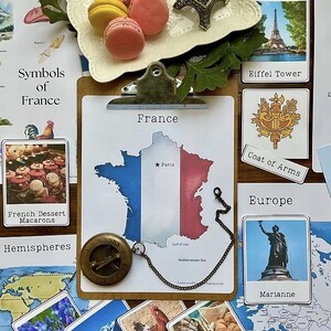 Könnte beinhalten: Eine Zusammenstellung französischer Themen, darunter eine Frankreichkarte, ein Miniatur-Eiffelturm und Karten mit Bildern von Macarons, dem Eiffelturm und dem französischen Wappen. Ein Kompass und ein Teller mit Macarons sind ebenfalls vorhanden.