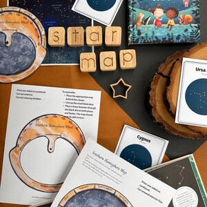 May include: A set of educational materials for learning about constellations. The set includes a book titled "What are Stars?" a wooden star map, and several constellation cards. The cards are printed on cardstock and feature illustrations of constellations, including Ursa Major, Cygnus, and Sagittarius. The cards are designed to be used with a viewer, which is not pictured.