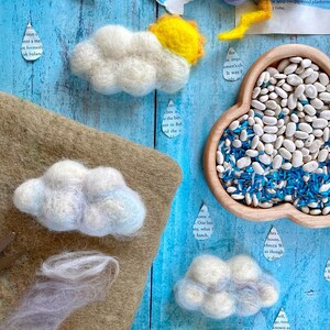 May include: A blue wooden background with white, gray, and blue felt clouds, a yellow felt sun, and a bowl of white beans and blue rice. The clouds are arranged in a line across the top of the image, with the sun in the center. The bowl of beans and rice is in the lower right corner of the image.