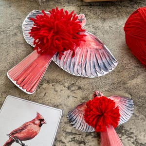 May include: Two red cardinal paper cutouts with red yarn pom-poms glued on top. The cardinals are in a flying position with their wings spread out. There is a smaller cardinal cutout in the bottom right corner of the image.