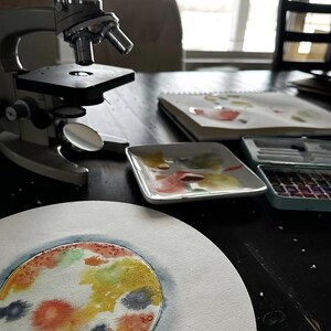 May include: A watercolor painting of a petri dish with colorful bacteria colonies. The painting is on a white canvas and is sitting on a black table. A microscope and a watercolor palette are also on the table.