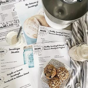 May include: An overhead shot of a baking scene with recipe sheets, a stand mixer, and baked muffins. The recipe sheets include titles like "The Science of Sourdough" and "Healthy Sourdough Discard Banana Breakfast Muffins." A small jar of starter and a bowl of oats are also visible.