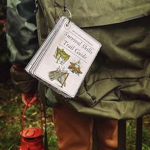 Pode incluir: Um conjunto de cartões brancos com o texto "Survival Skills Trail Guide" está preso a uma mochila. O guia apresenta ilustrações de uma tenda, uma fogueira e um abrigo. Uma lanterna vermelha é segurada na mão, e a pessoa está vestindo uma jaqueta verde e calças marrons.