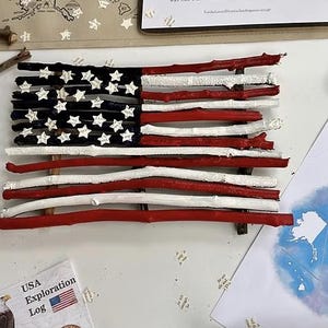 May include: A handcrafted American flag made from painted sticks. The flag features red and white stripes and a blue canton with white stars. Additional elements include a map of the USA, a USA Exploration Log, and star-shaped confetti.