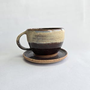 May include: A handcrafted ceramic coffee cup and saucer. The cup has a two-tone design, with a light beige upper section and a dark brown lower section. The saucer is a matching dark brown. Perfect for serving tea or coffee.