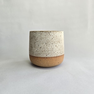 May include: A ceramic tumbler with a speckled design and a rounded base. The upper section is light beige with dark speckles, and the lower section is a warm brown. The cup has a smooth, unadorned surface.