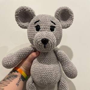 Könnte beinhalten: Ein handgefertigtes graues Plüschmaus-Spielzeug mit großen Ohren, schwarzen Augen und einer schwarzen Nase. Das Stofftier besteht aus einem strukturierten Material und hat Arme und Beine. Die Maus ist ein niedliches, kuscheliges Design.