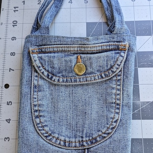 May include: A small blue denim bag with a single pocket and a gold button. The bag has two straps that can be worn over the shoulder.