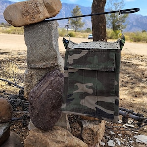 May include: A camouflage fabric crossbody bag with a white lace trim and a zippered pocket. The bag is hanging from a metal hook.