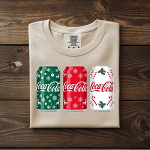 May include: A beige Comfort Colors t-shirt with three Coca-Cola cans featuring holiday designs. The cans are green with snowflakes, red with snowflakes, and white with candy canes and holly. The shirt is folded on a wooden surface.