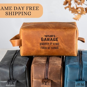 May include: A rectangular leather toiletry bag in tan, black, brown, and blue. The tan bag features the text 'TAYLOR'S GARAGE KNOWER OF STUFF FIXER OF THINGS'. Each bag has a side handle and a zipper closure with a small leather tab showing initials.