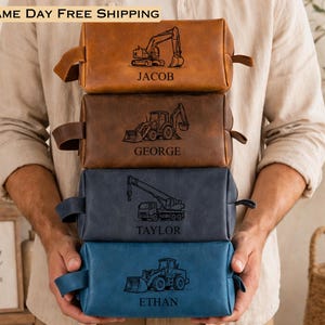 May include: A stack of four rectangular leather toiletry bags in tan, brown, dark gray, and blue. Each bag features a laser-engraved construction vehicle graphic and a name: Jacob with an excavator, George with a backhoe, Taylor with a crane, and Ethan with a loader.